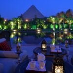 From above of cafe with dishware and ornamental lanterns on round tables near sofa and pond reflecting trees and old pyramid under blue sky illuminated by artificial lights
